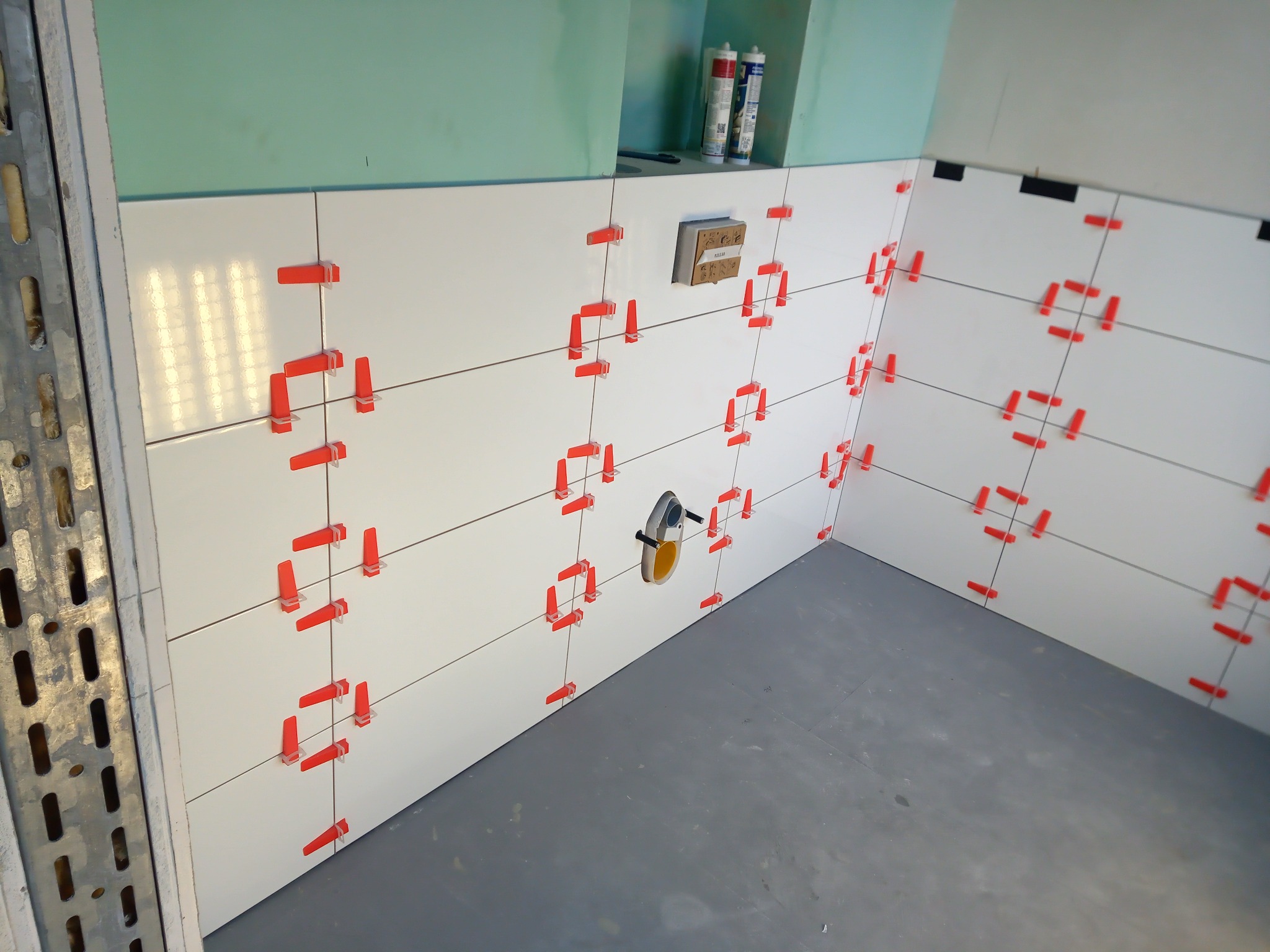 Świeżo ułożone, białe płytki ścienne z czerwonymi klipsami poziomującymi, widoczne przewody elektryczne, surowy stan ściany i podłogi, przygotowanie do wykończenia łazienki.