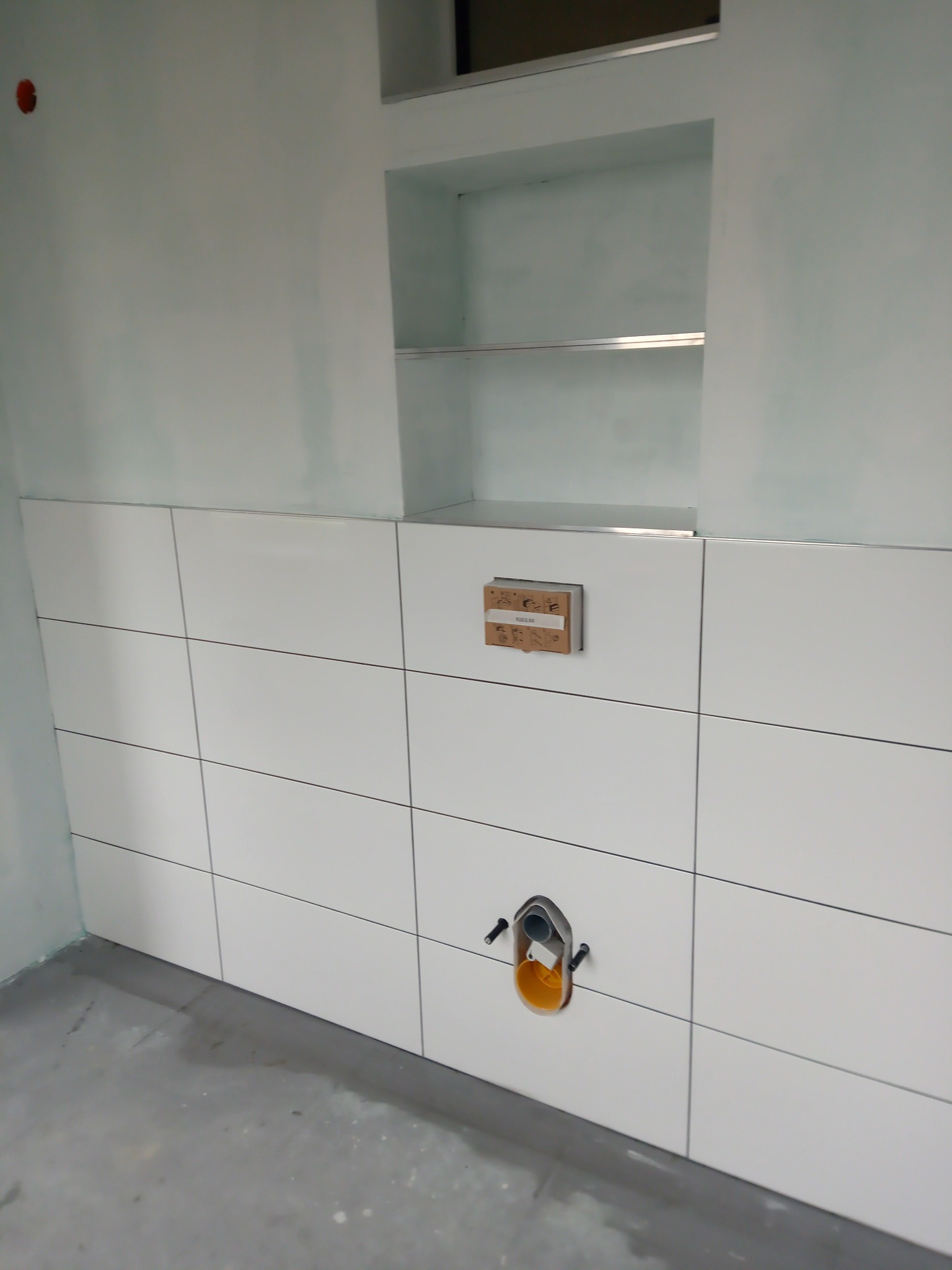 Ściana w trakcie wykańczania z białymi, kwadratowymi płytkami ceramicznymi, wnęka z półkami, instalacja podtynkowy WC z żółtym elementem, surowy beton na podłodze i ślady po pracach wykończeniowych...