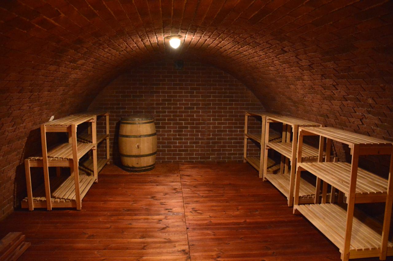 Piwnica z ceglanym sklepieniem łukowym, drewnianą podłogą, beczką i surowymi, sosnowymi regałami po bokach.