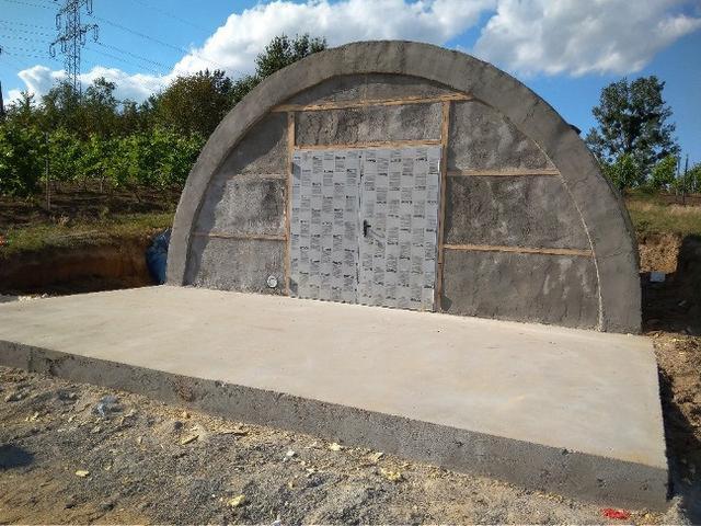 Betonowa konstrukcja w kształcie łuku z drewnianymi elementami i zabezpieczonymi drzwiami, w otoczeniu zieleni i ziemi.