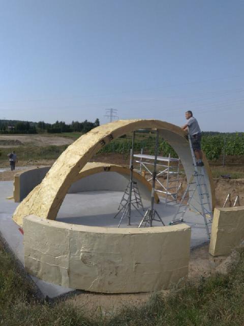 Budowa nietypowej altany z łukowatą konstrukcją drewnianą, widoczny pracownik na drabinie i częściowo wykończone betonowe fundamenty.