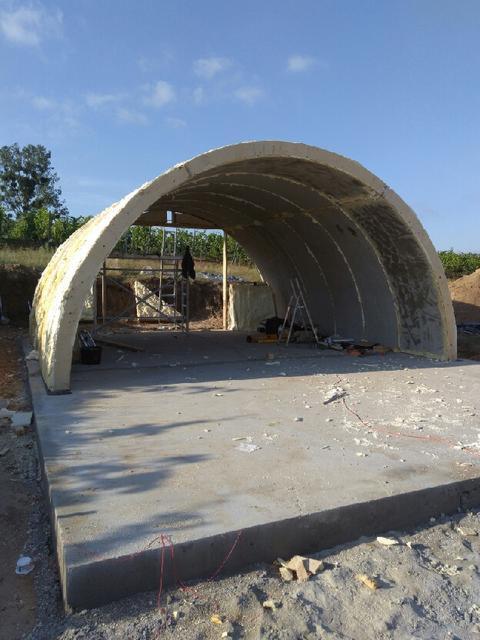 Konstrukcja łukowa z betonu na fundamencie, w trakcie budowy, z widocznymi rusztowaniami i narzędziami w tle, otoczona zielenią i błękitnym niebem.