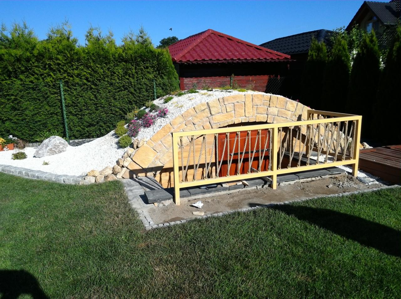 Ziemianka ogrodowa z kamienną fasadą, białym żwirem i żółtą balustradą na trawniku w słoneczny dzień.