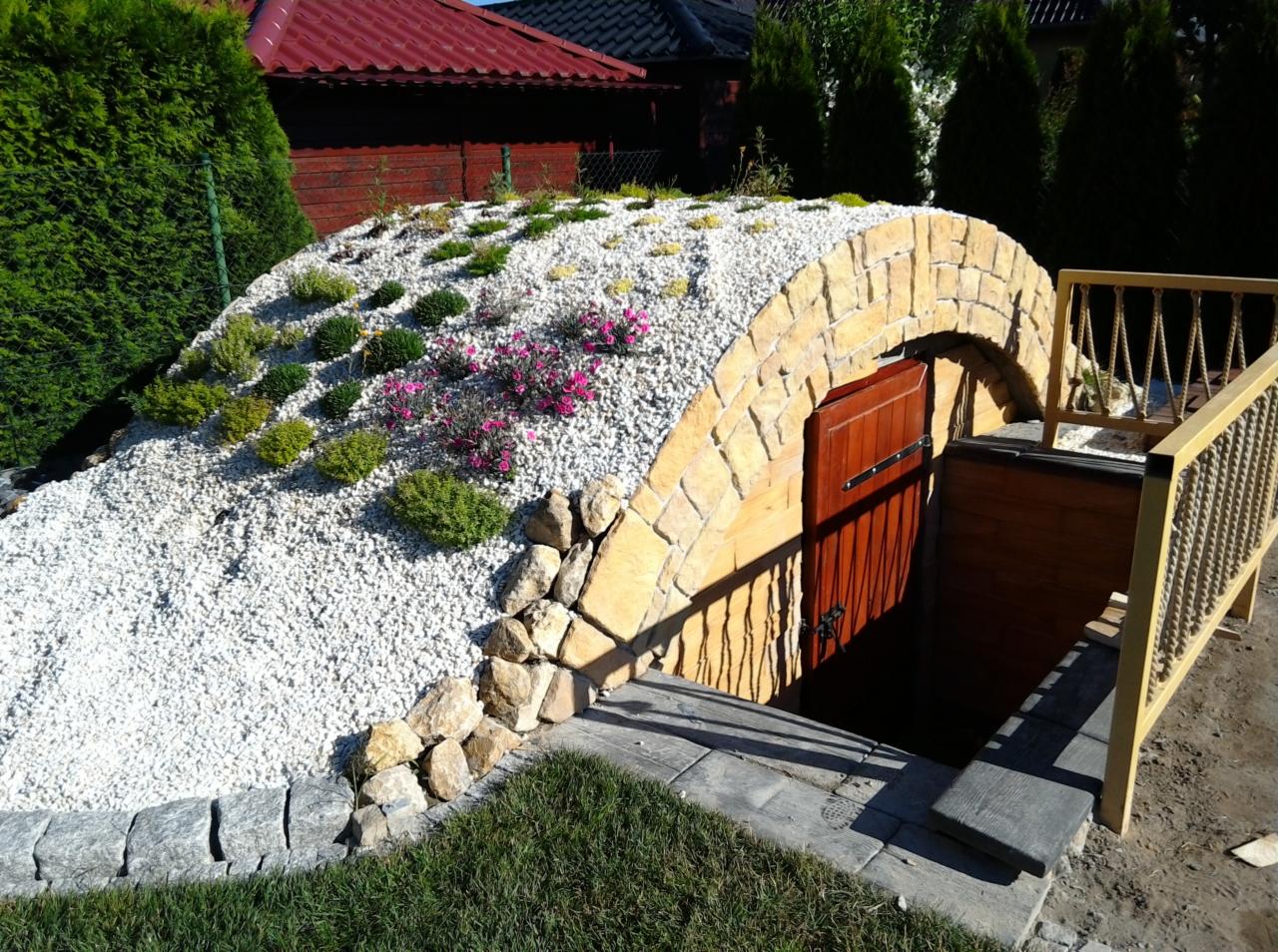 Ziemianka z kamienną elewacją, drewnianymi drzwiami i schodami, obsypana białym żwirem i obsadzona roślinami, widok z zewnątrz.