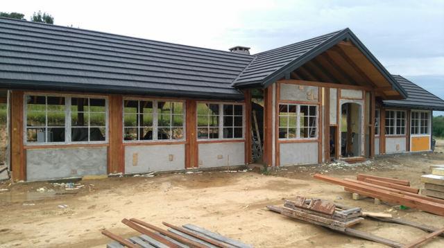 Budynek w trakcie rozbudowy z widoczną konstrukcją drewnianą, oknami z białymi ramami i szarą dachówką, z elementami ocieplenia i surowym otoczeniem budowy.