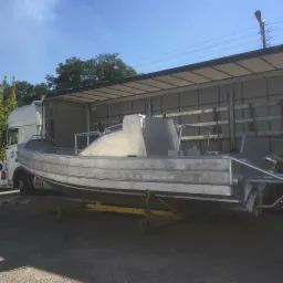 Surowy, aluminiowy kadłub łodzi na ż&oacute;łtym w&oacute;zku transportowym, częściowo załadowany na naczepę ciężar&oacute;wki w słoneczny dzień.