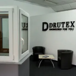 Ekspozycja dwuskrzydłowego okna z białymi ramami, otwartego w pomieszczeniu z logo firmy Drutex na ścianie, widoczne krzesła i stolik.