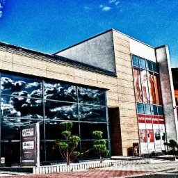 Budynek biurowy w Trzebini z dużymi oknami, w których odbija się błękitne niebo z chmurami. Przed wejściem stoją tablice reklamowe i ozdobne drzewka.