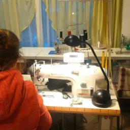 Kobieta w pomarańczowej bluzie siedzi przy białej maszynie do szycia marki Siruba, oświetlonej lampką; w tle okno z żółtymi zasłonami i inne stanowiska pracy.