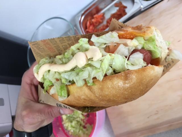 Ręka trzymająca kanapkę z długą bułką, sałatą, sosem i dodatkami, zapakowaną w papier z nadrukiem.