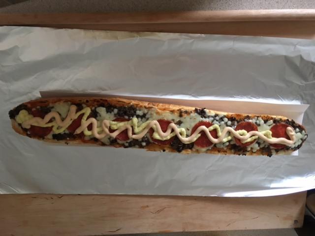 Długa kanapka na bagietce z pepperoni, serem, brązowym farszem i sosem, ułożona na folii aluminiowej.