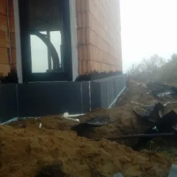 Ocieplenie fundamentów budynku z bloczków, widoczne czarne płyty izolacyjne, wykop ziemny i instalacje na zewnątrz.