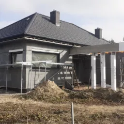 Surowy dom jednorodzinny w trakcie budowy, ściany z szarego bloczka, częściowo ocieplone białym styropianem, widoczne rusztowania i sterty ziemi.