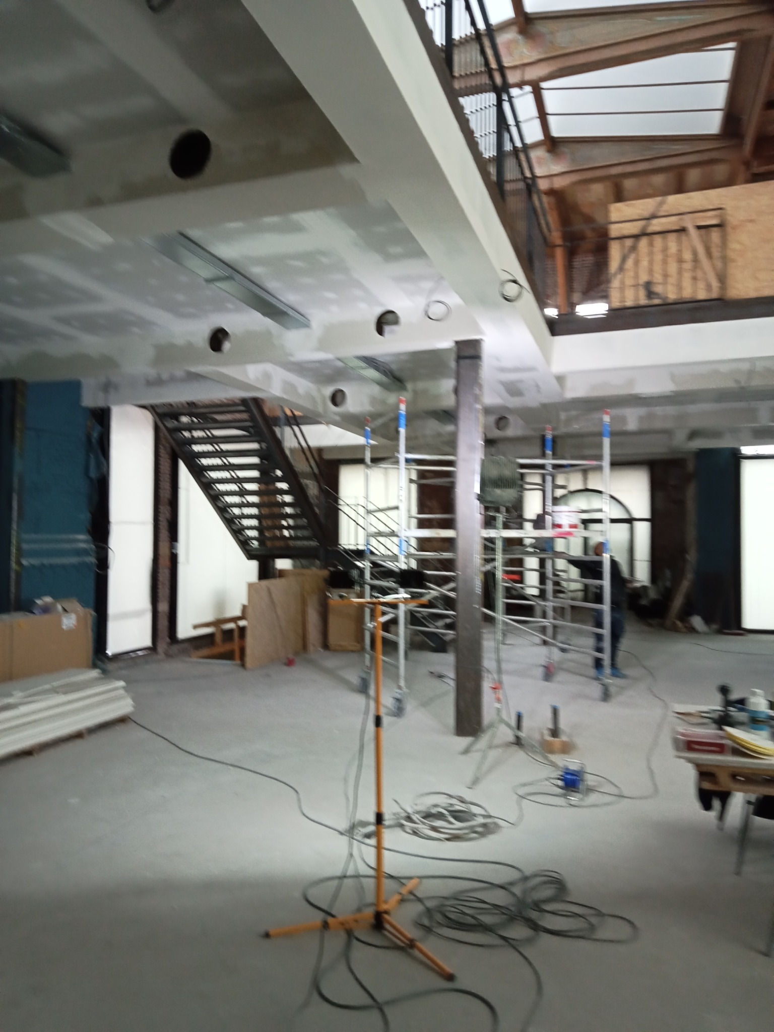 Wnętrze w trakcie remontu: widoczne rusztowania, przewody elektryczne na podłodze, częściowo wykończony sufit z otworami na oświetlenie, oraz metalowe schody w tle.