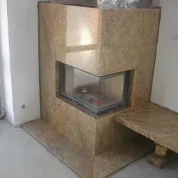 Obudowa kominka z jasnego, ziarnistego kamienia z przeszklonym trójkątnym paleniskiem, widoczny fragment podłogi w trakcie montażu.