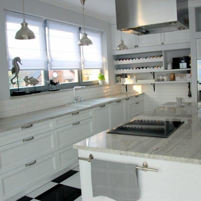 Jasna kuchnia z białymi szafkami, blatem z jasnego kamienia z delikatnym wzorem, czarno-białą podłogą w szachownicę i dekoracjami okiennymi z motywami morskimi.