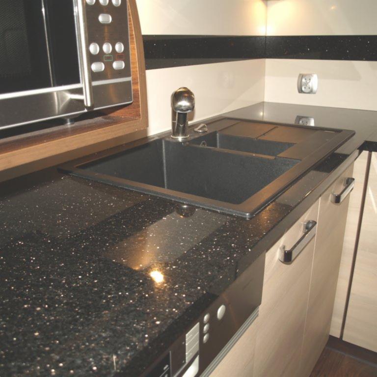 Czarny, błyszczący blat kuchenny z wbudowanym, czarnym, prostokątnym zlewem i chromowaną baterią, widoczny fragment zabudowy kuchennej z jasnymi frontami i stalowymi uchwytami.