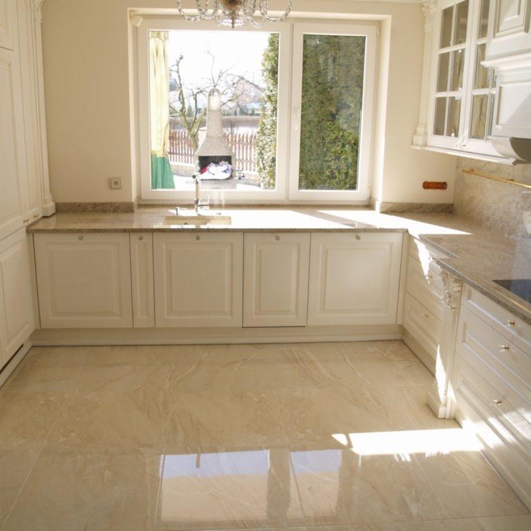 Jasna kuchnia z beżowymi płytkami podłogowymi, białymi szafkami z frezowanymi frontami i blatem z kamienia o podobnej kolorystyce; widok na zewnątrz przez okno z grillem ogrodowym w tle.