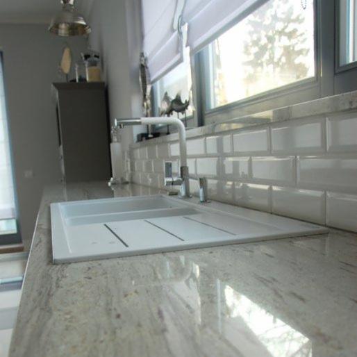 Jasny, polerowany blat kuchenny z granitu z wbudowanym białym zlewozmywakiem i baterią, tło stanowią białe, prostokątne płytki ułożone na ścianie pod oknem.