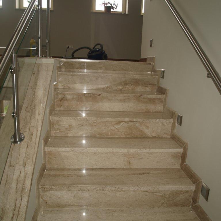 Jasne, beżowe schody wewnętrzne wykończone kamieniem z metalową balustradą i szklanym wypełnieniem, widoczny odkurzacz w tle.
