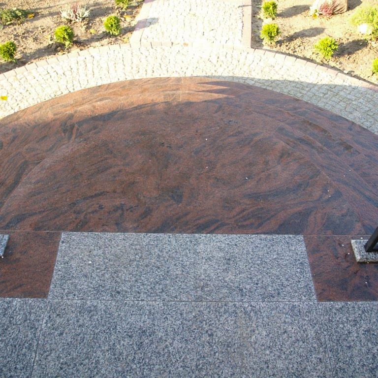 Granitowy podest z dwoma stopniami w odcieniach szarości i czerwieni, otoczony kostką brukową i zielenią.