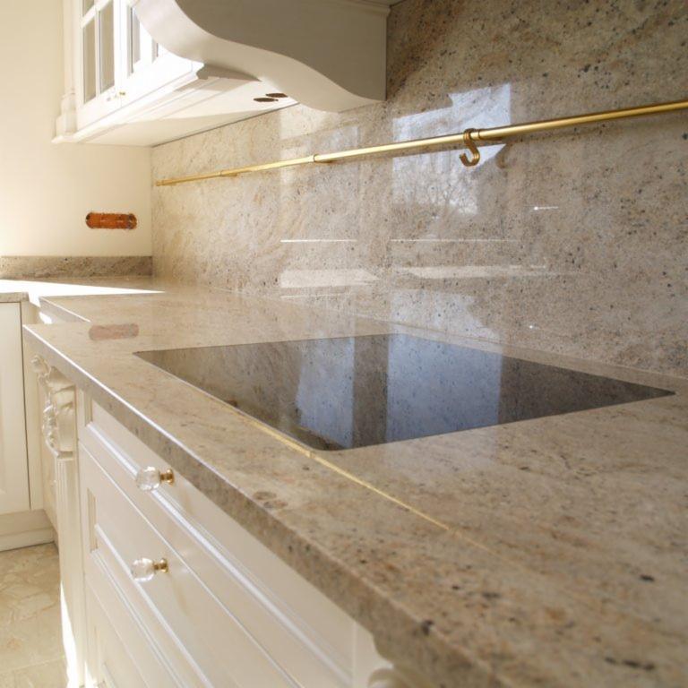 Jasny blat kuchenny z kamienia naturalnego z wbudowanym zlewem, kremowe szafki z ozdobnymi uchwytami, złoty reling na ścianie.