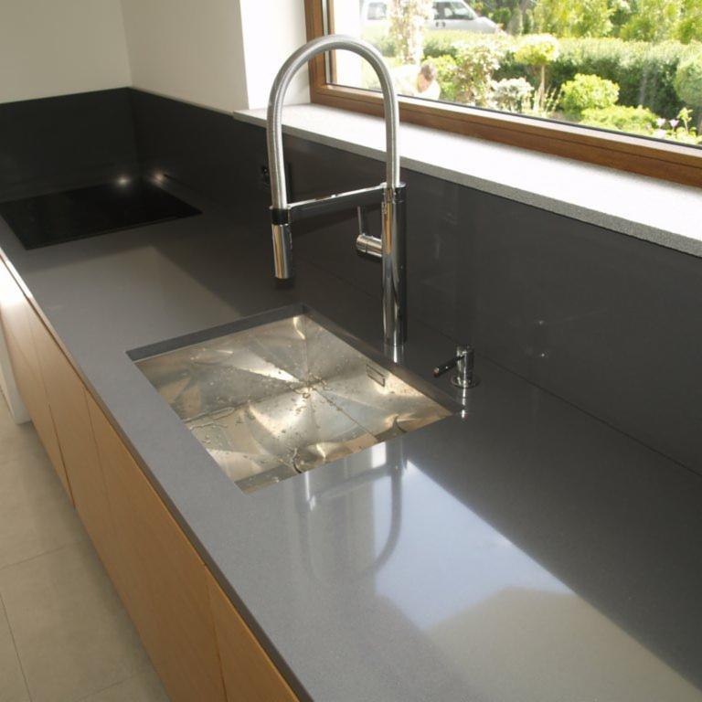 Szary blat kuchenny z metalowym zlewem i chromowaną baterią na tle okna z widokiem na zieleń.