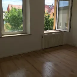 Pusty pokój z dwoma oknami, jeden z łukowym nadprożem, podłoga z paneli, białe ściany i grzejnik pod oknem.