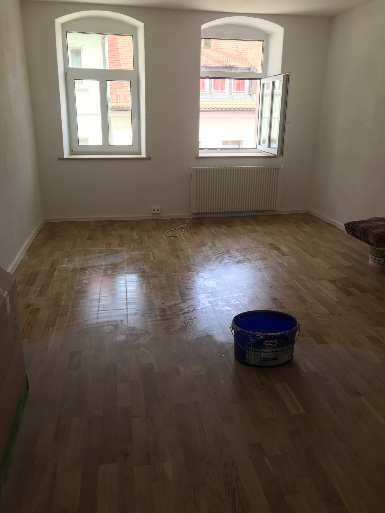 Pokój po malowaniu ścian, widoczne dwa okna, grzejnik, drewniana podłoga z plamami farby i niebieskie wiadro z farbą przy podłodze.