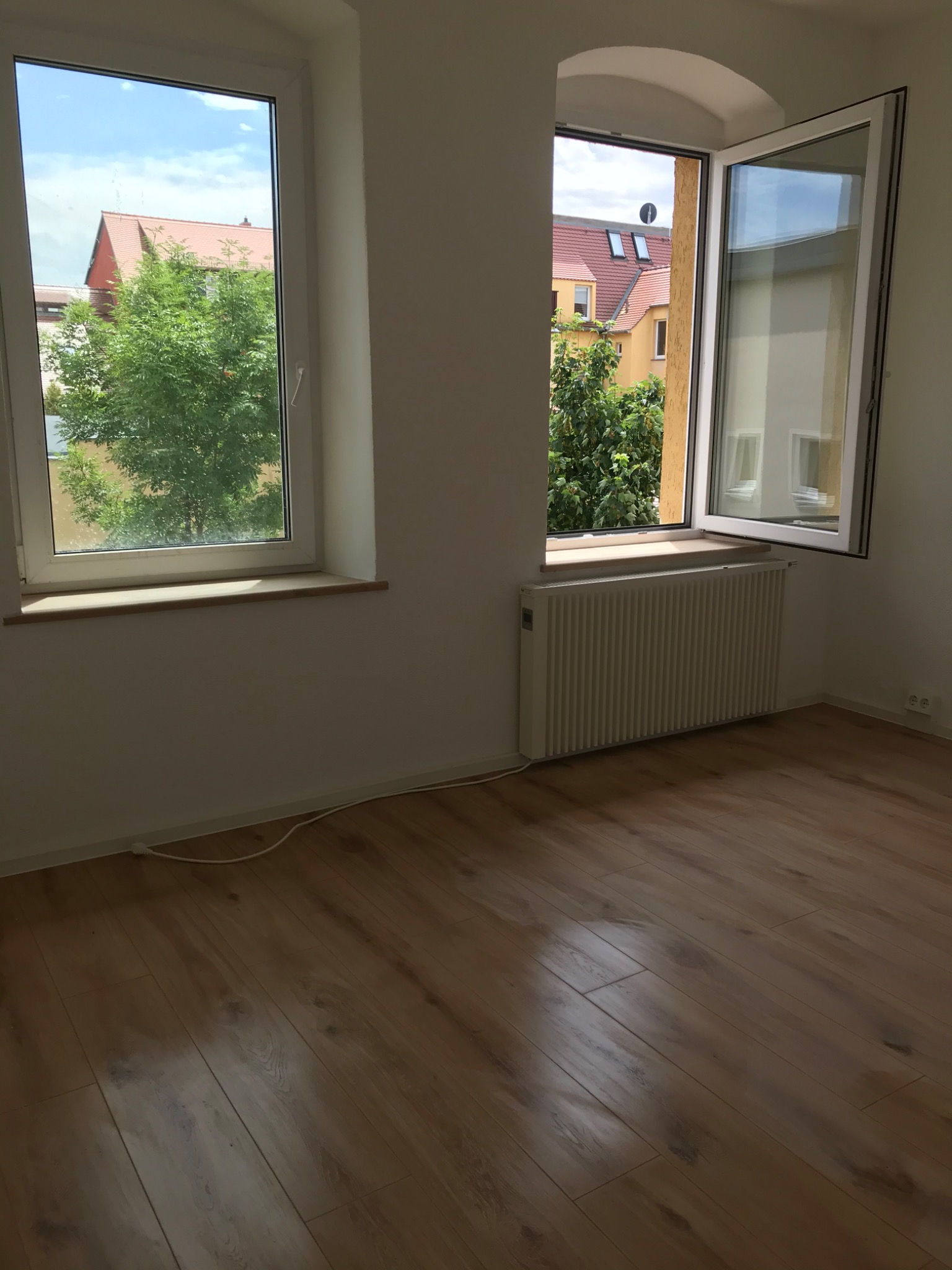 Pusty pokój z dwoma oknami, jeden z łukowym nadprożem, podłoga z paneli, białe ściany i grzejnik pod oknem.