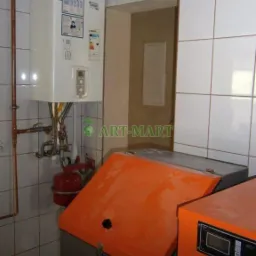 Kocioł grzewczy z podajnikiem na ekogroszek w kotłowni z białymi kafelkami, widoczny biały piec gazowy na ścianie i instalacja rur.