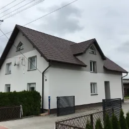 Dwupiętrowy dom jednorodzinny z brązowym dachem, świeżo otynkowaną białą elewacją i numerem 16, widziany z poziomu chodnika w pochmurny dzień.