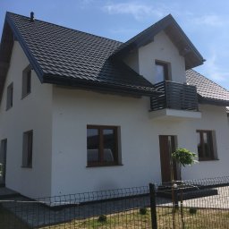 Zakład Ogólnobudowlany ZEN- BUD Zenon Kuźnik - Nowoczesny dom jednorodzinny z białą elewacją, ciemną dachówką i drewnianymi oknami, widok z niskiej perspektywy przez ogrodzenie.