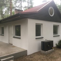 Zakład Ogólnobudowlany ZEN- BUD Zenon Kuźnik - Biały, otynkowany domek jednorodzinny z ciemną, drewnianą elewacją szczytową i okrągłym oknem, otoczony zielenią, z widocznym agregatem klimatyzacji.