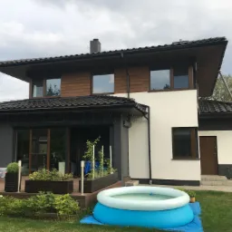 Nowoczesny dom dwukondygnacyjny z drewnianą elewacją na piętrze, jasną elewacją na parterze, tarasem z meblami ogrodowymi i basenem dmuchanym na trawniku.