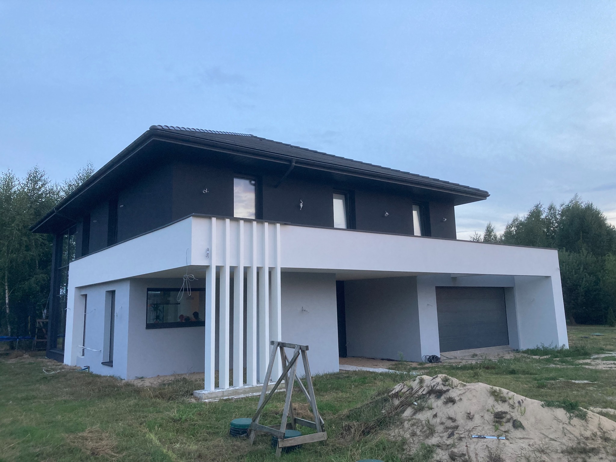 Nowoczesny dom jednorodzinny z ciemną elewacją i białymi elementami architektonicznymi, widoczny z zewnątrz, z drewnianą kozą malarską na trawniku przed budynkiem.