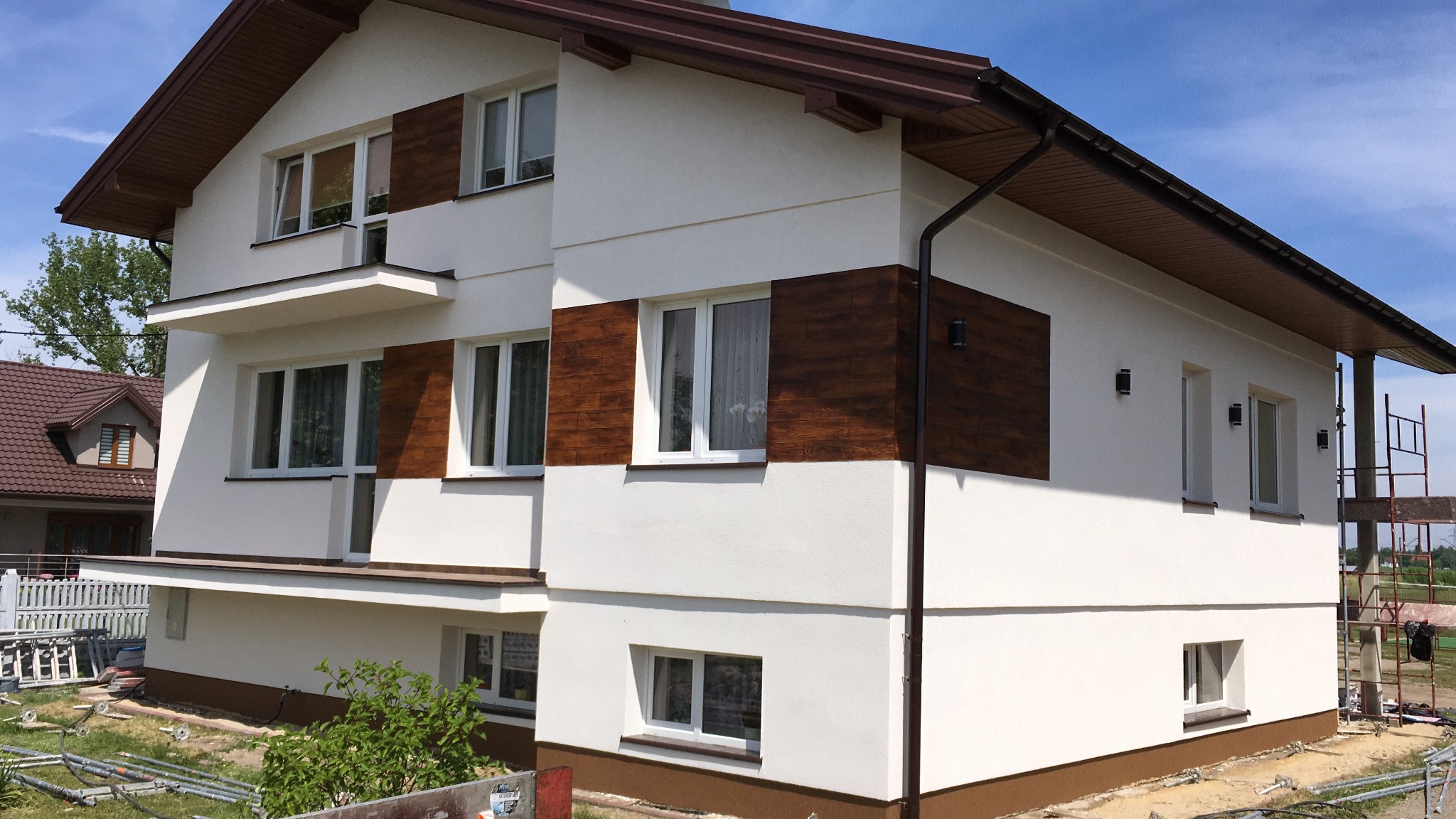 Dwupiętrowy dom z białą elewacją i brązowymi panelami dekoracyjnymi, widoczne rusztowania z boku budynku.