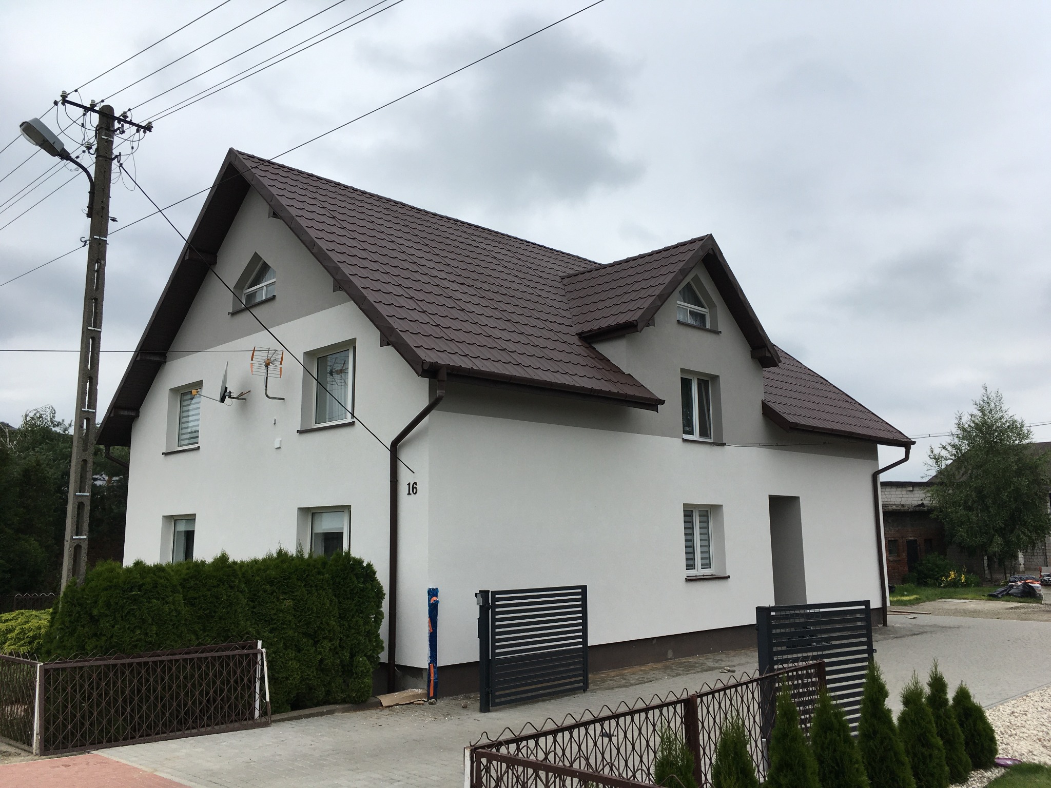 Dwupiętrowy dom jednorodzinny z brązowym dachem, świeżo otynkowaną białą elewacją i numerem 16, widziany z poziomu chodnika w pochmurny dzień.