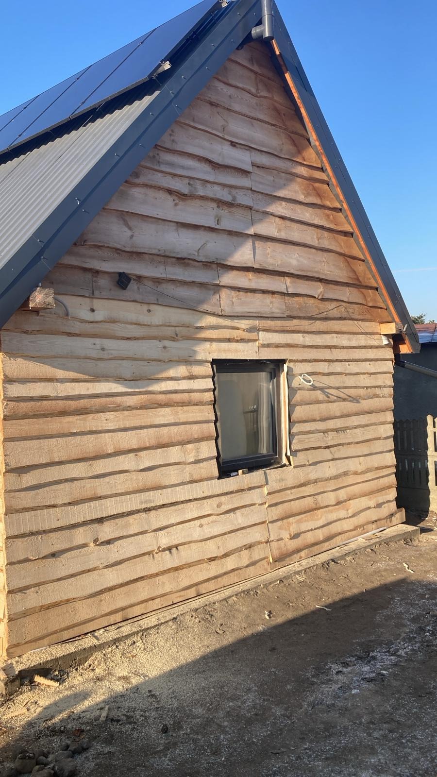 Surowa drewniana elewacja domu jednorodzinnego z zamontowanym oknem i panelami słonecznymi na dachu, widok z dołu w słoneczny dzień.