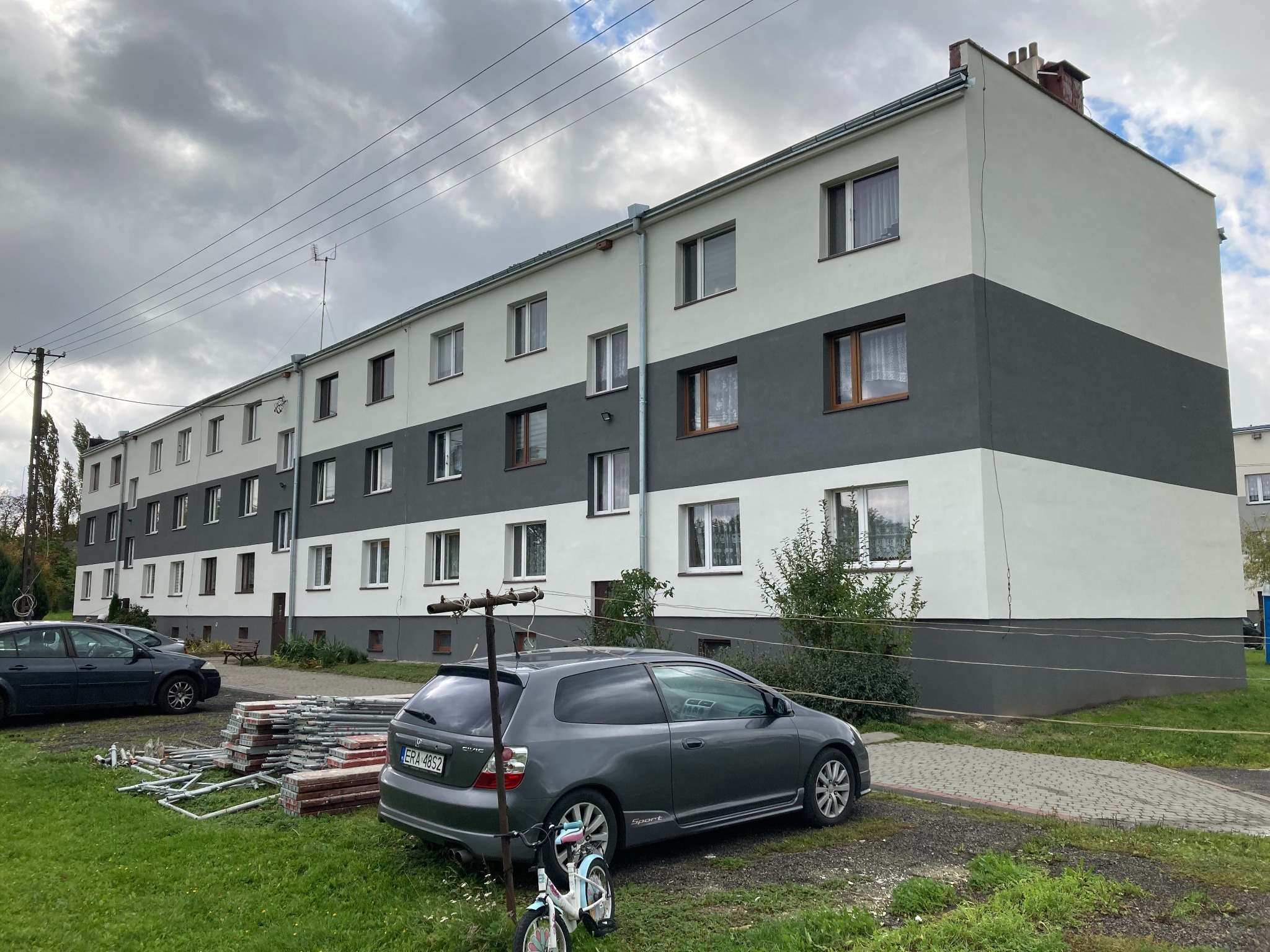 Wielorodzinny budynek po renowacji elewacji z dwukolorowym malowaniem (biały i szary), na trawniku zaparkowany szary samochód osobowy i dziecięcy rowerek, obok sterta płyt chodnikowych i rozłożone...