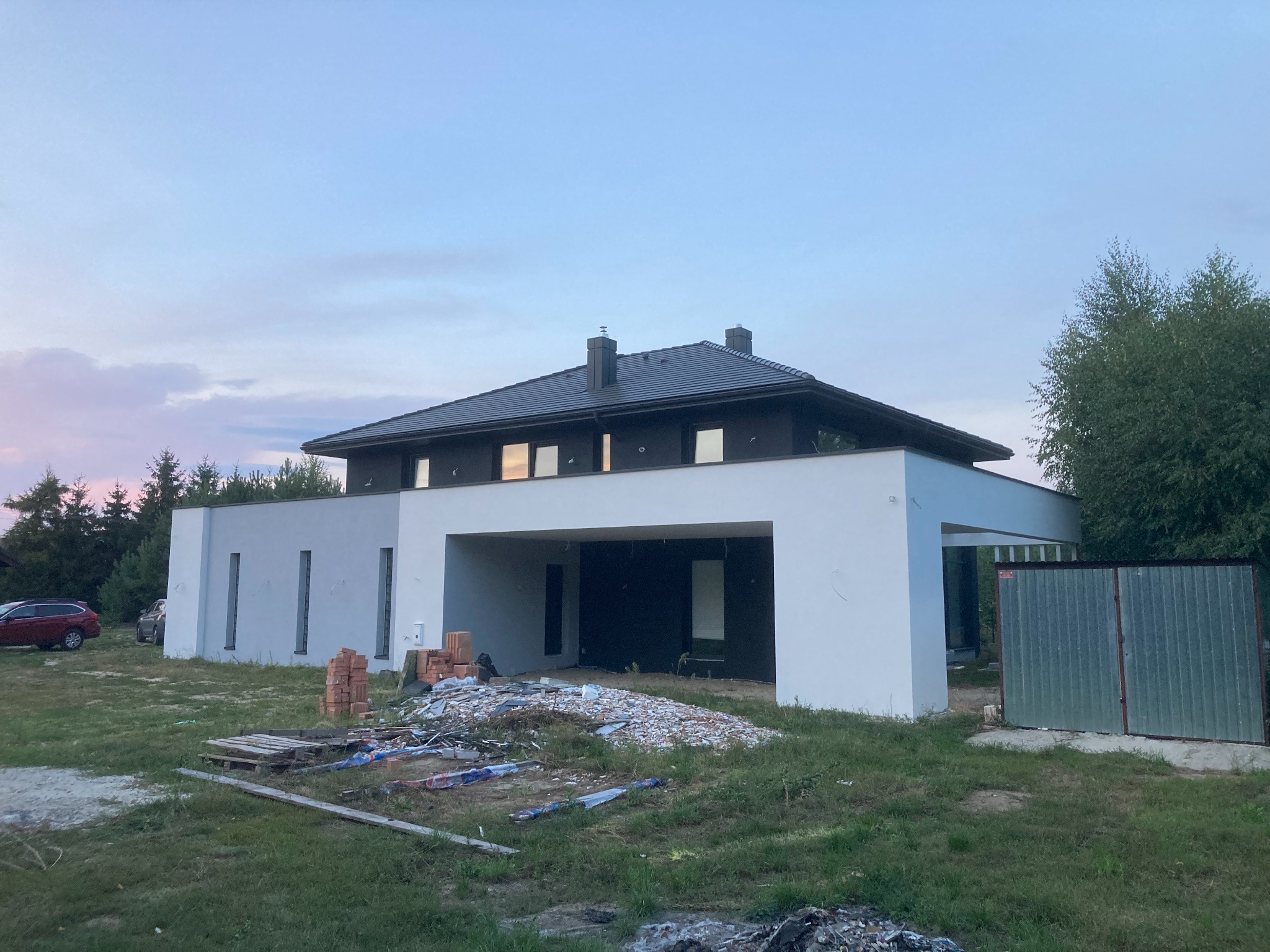 Surowy stan elewacji nowoczesnego domu jednorodzinnego z widocznymi niedokończonymi pracami, stertą gruzu i cegieł na trawniku przed budynkiem.