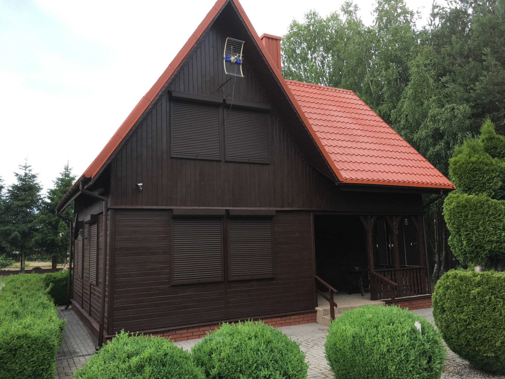 Brązowy, drewniany dom z czerwoną dachówką i zewnętrznymi roletami, otoczony zielonymi krzewami i drzewami. Na dachu widoczna antena.