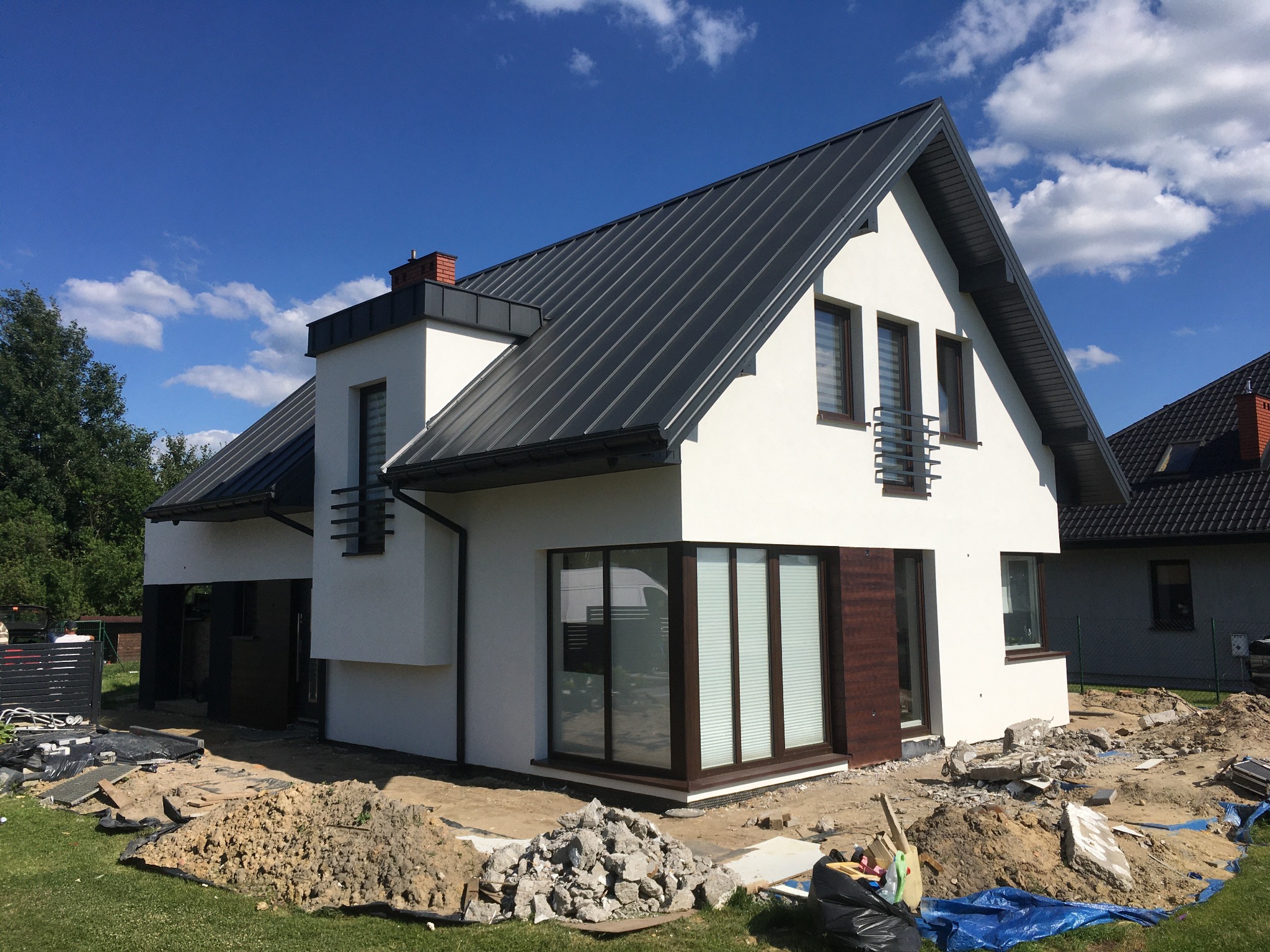 Nowoczesny dom jednorodzinny z białą elewacją i grafitowym dachem, widoczny w trakcie prac wykończeniowych wokół budynku, z hałdą ziemi i gruzu na trawniku.