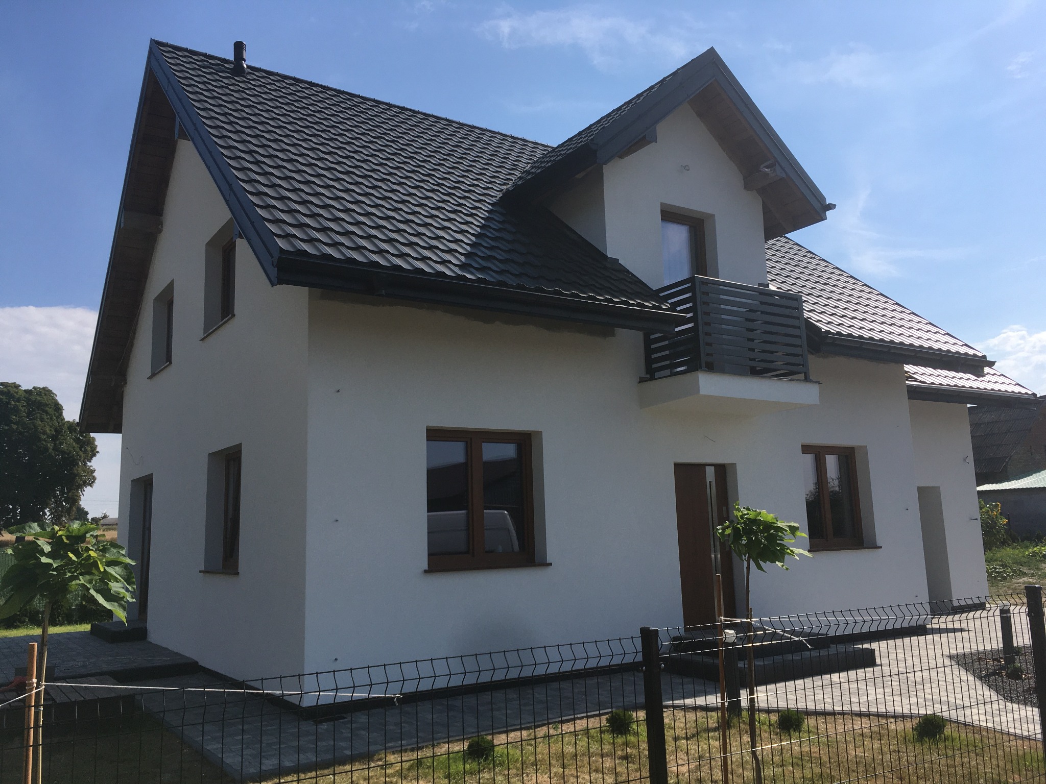 Nowoczesny dom jednorodzinny z białą elewacją, ciemną dachówką i drewnianymi oknami, widok z niskiej perspektywy przez ogrodzenie.