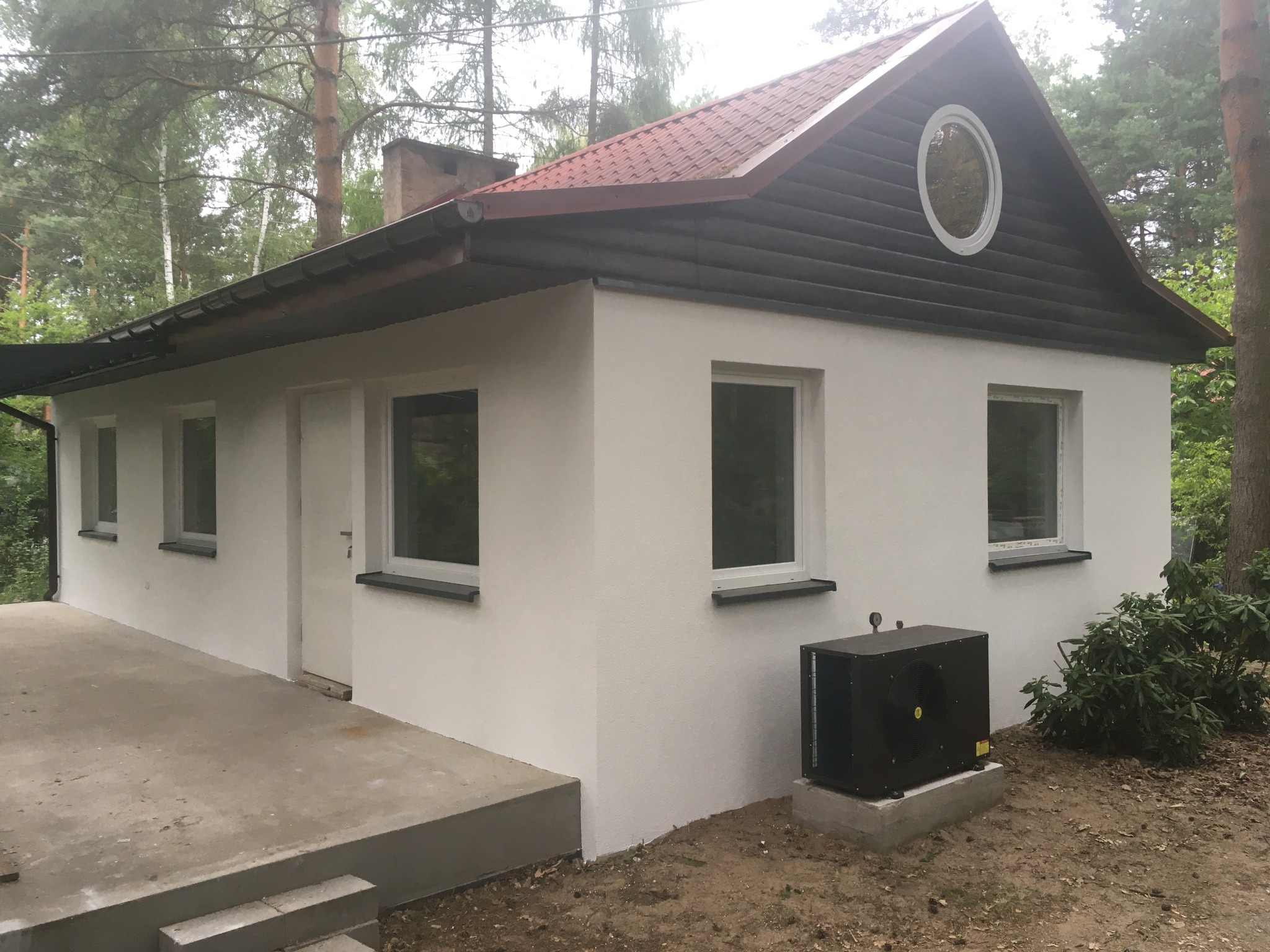 Biały, otynkowany domek jednorodzinny z ciemną, drewnianą elewacją szczytową i okrągłym oknem, otoczony zielenią, z widocznym agregatem klimatyzacji.