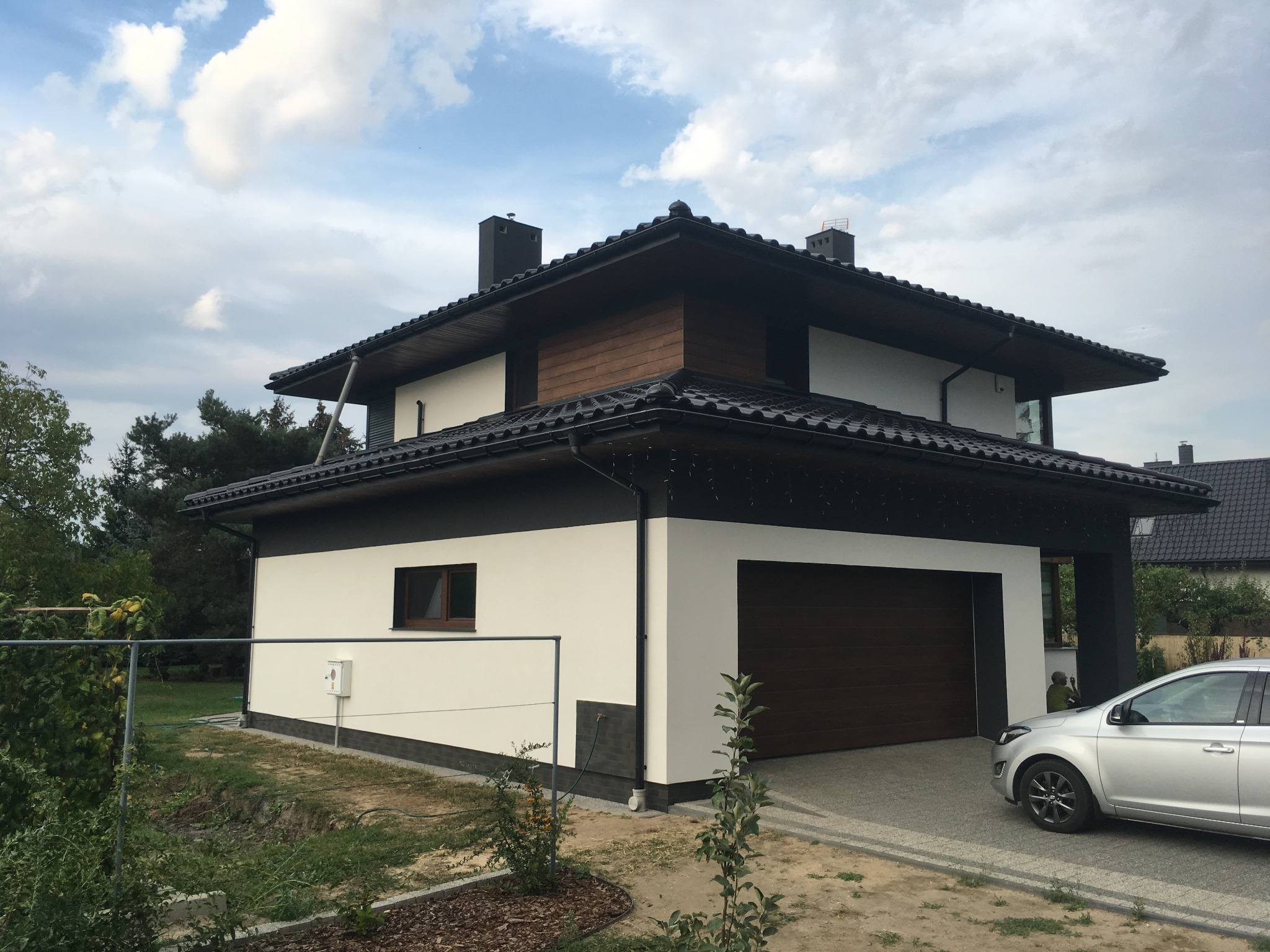 Nowoczesny dom jednorodzinny z białą elewacją, ciemnym dachem i drewnianymi akcentami, widoczny z boku, z samochodem zaparkowanym na podjeździe.