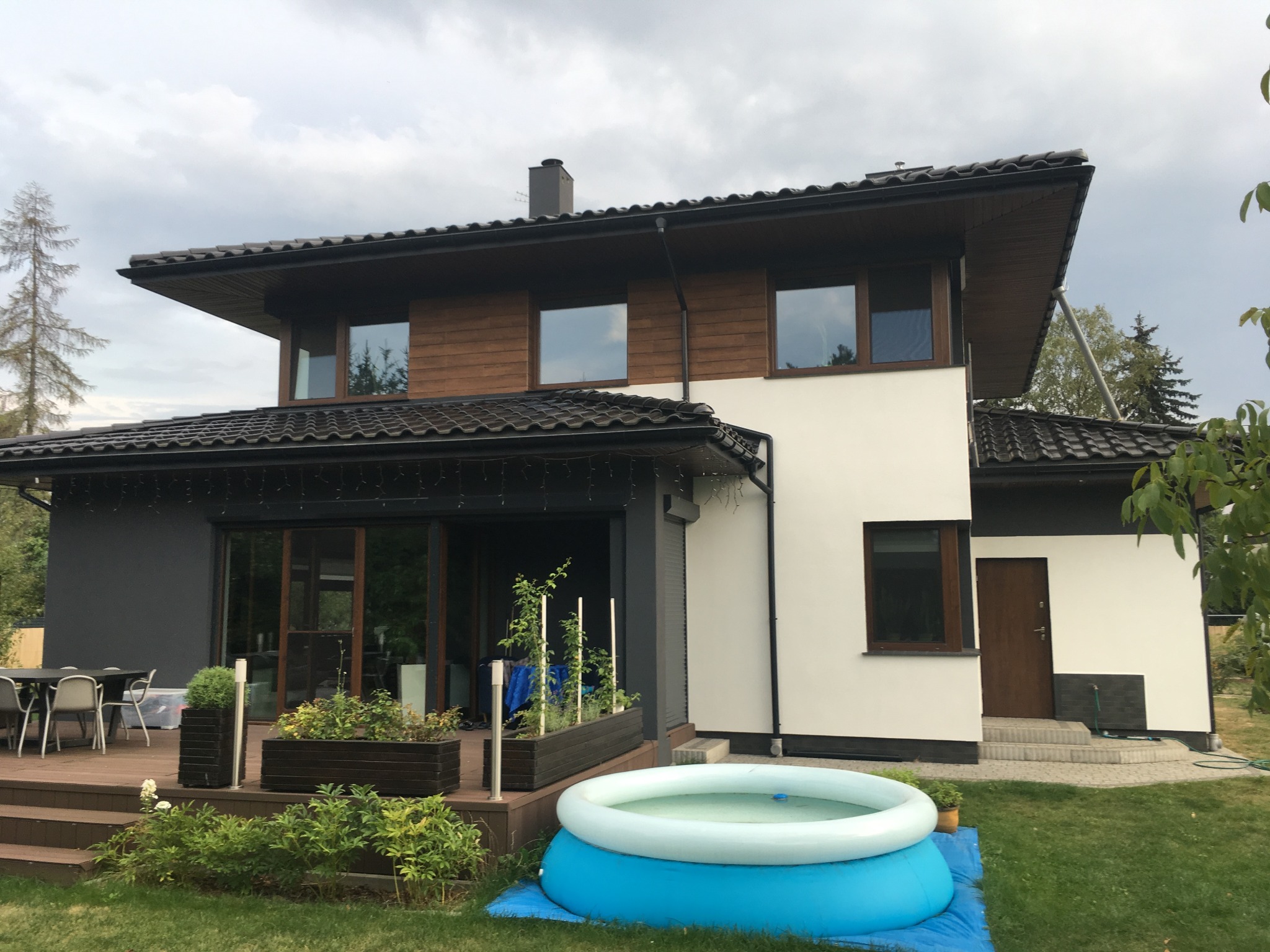 Nowoczesny dom dwukondygnacyjny z drewnianą elewacją na piętrze, jasną elewacją na parterze, tarasem z meblami ogrodowymi i basenem dmuchanym na trawniku.