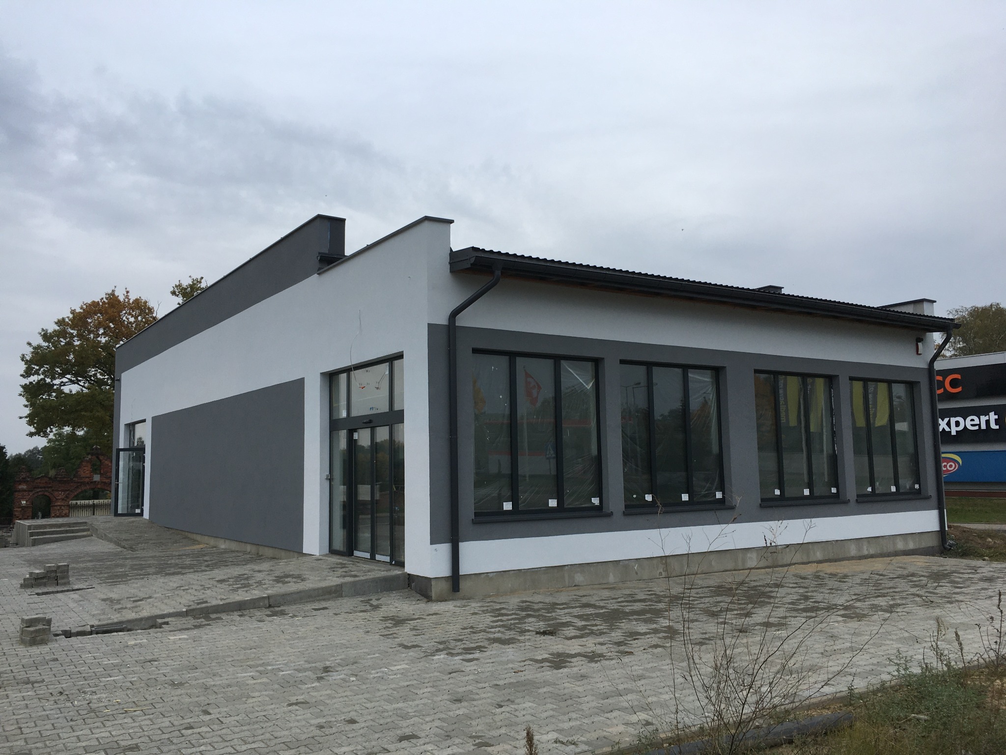 Nowoczesny budynek handlowy z szaro-białą elewacją, widoczne okna z czarnymi ramami i chodnik z kostki brukowej, pochmurne niebo w tle.