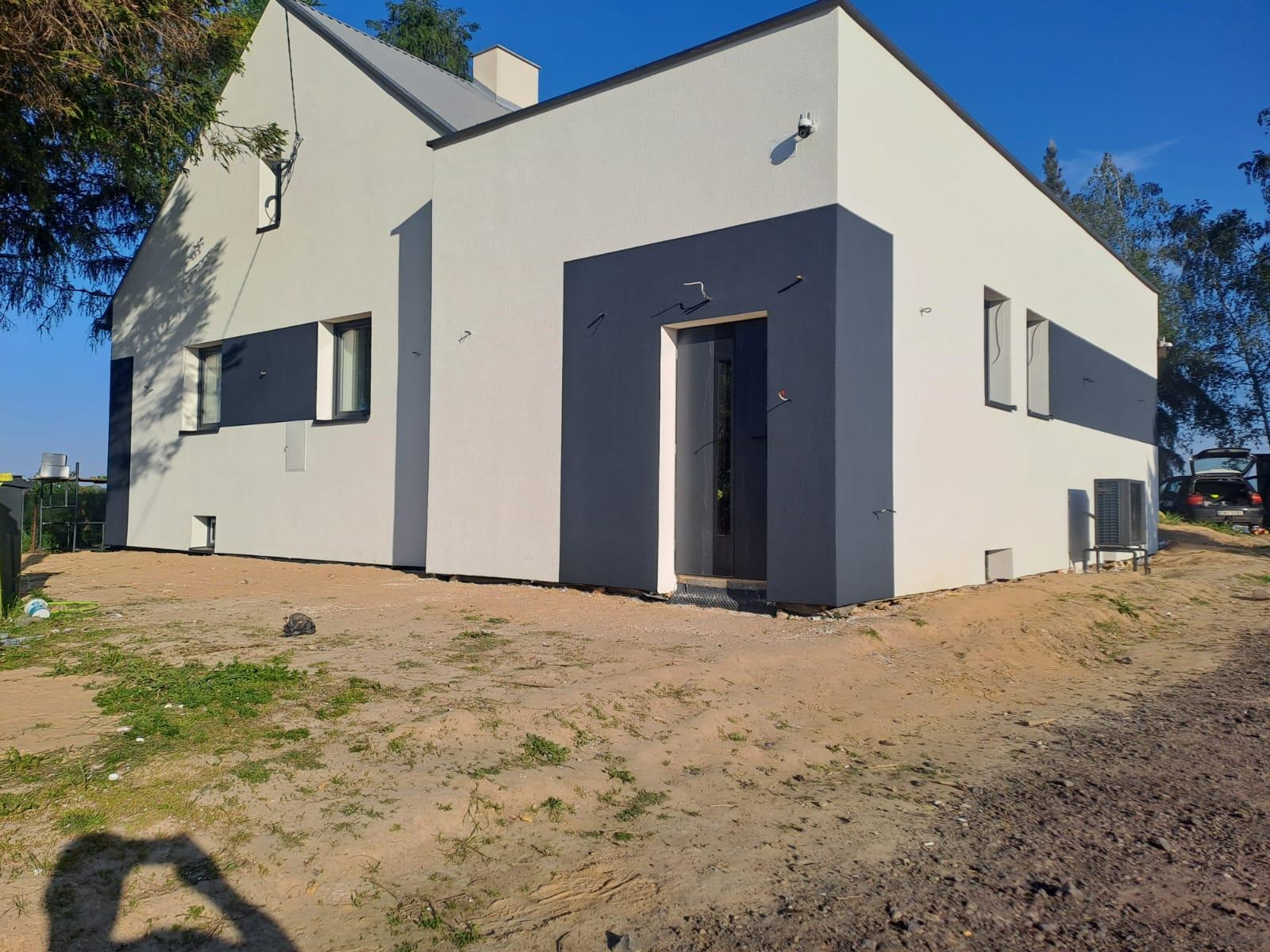 Nowoczesny dom jednorodzinny z biało-szarą elewacją, ciemnymi akcentami wokół okien i drzwi, widoczne pozostałości po pracach budowlanych na gruncie.