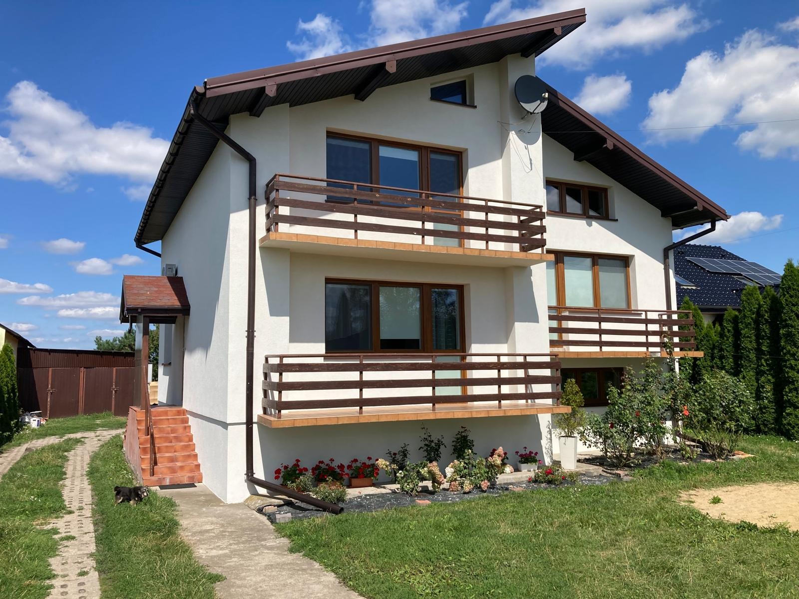 Dwupiętrowy dom z białym tynkiem, brązowymi ramami okiennymi i balustradami balkonowymi, widoczny z poziomu trawnika z kwiatami i małym psem.
